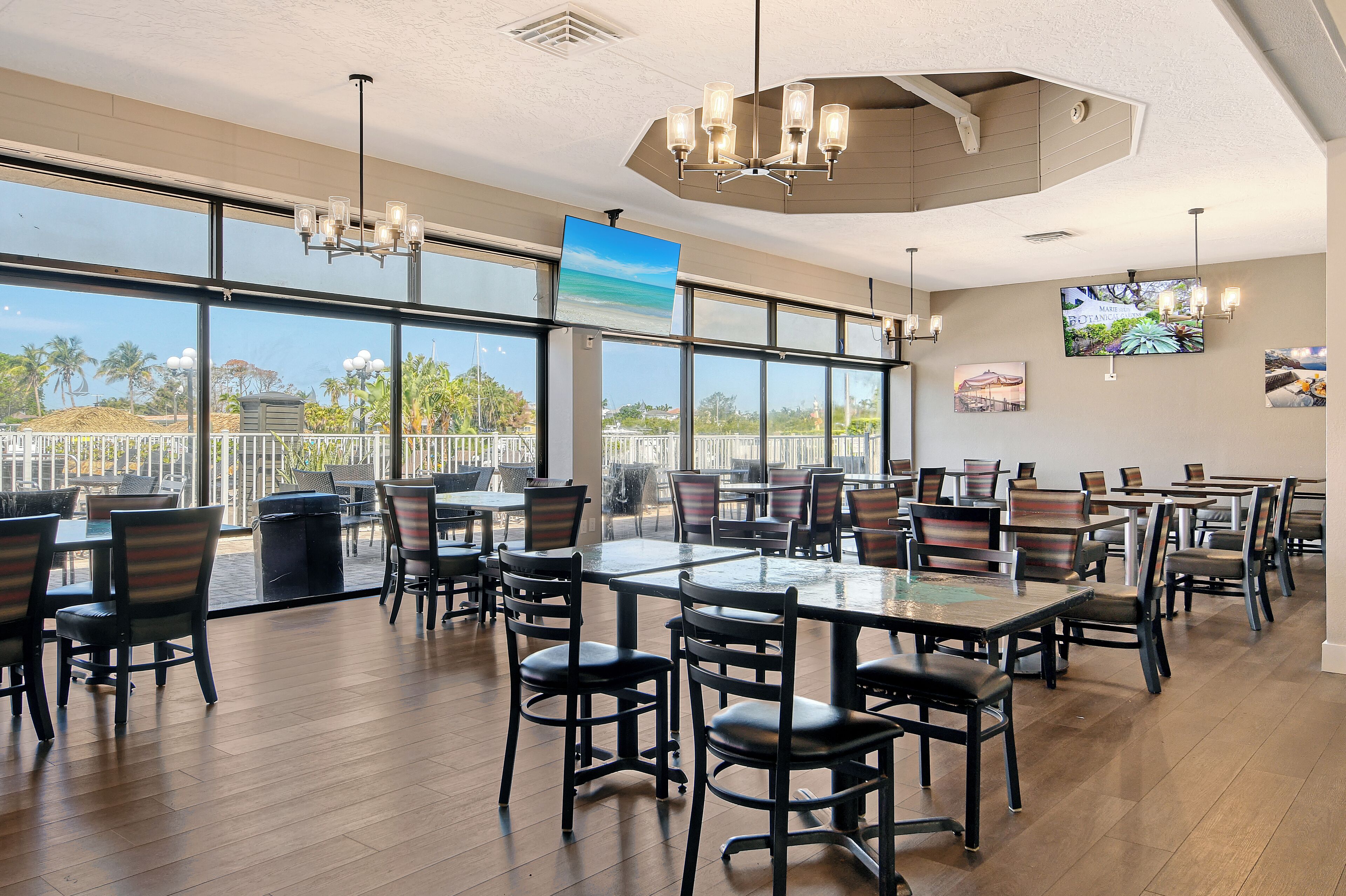 A spacious indoor dining area with multiple tables and chairs, large windows offering a view of palm trees and a beach, and a modern chandelier.