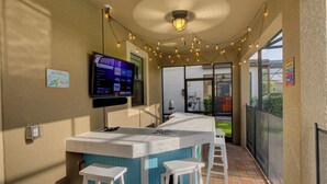 Un pequeño bar al aire libre con un televisor montado en la pared, luces colgantes y vista al jardín trasero.