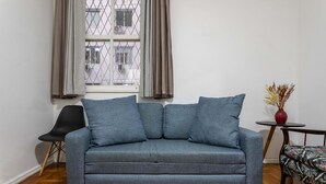 Ein graues Sofa mit vielen Kissen, ein schwarzer Sessel, ein kleiner runder Tisch mit einer Vase und ein Fenster mit Vorhängen.