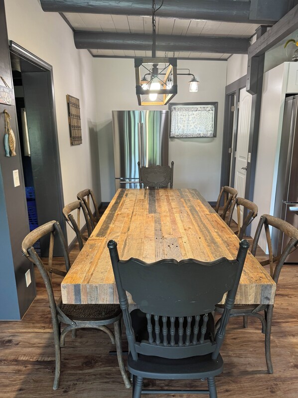A dining area with a long wooden table, six chairs, a chandelier, a refrigerator, and a patterned wall hanging.
