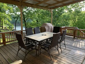 A covered outdoor deck with a table and chairs, overlooking a lake and surrounded by trees.