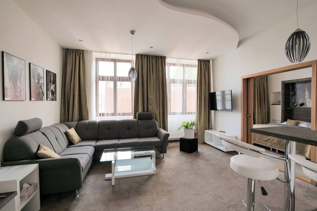 A modern living room with a sectional sofa, a glass coffee table, a dining area with a bar stool, and a TV mounted on the wall.