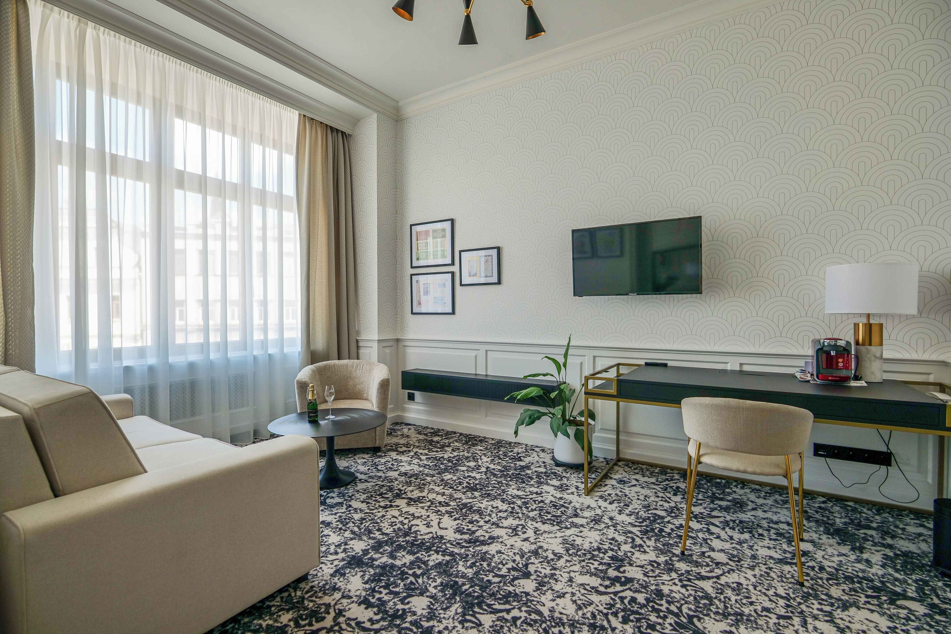 Ein modernes Hotelzimmer mit einer Couch, einem runden Couchtisch, einem Schreibtisch mit Lampe, einem Fernseher und gerahmten Bildern an der Wand.