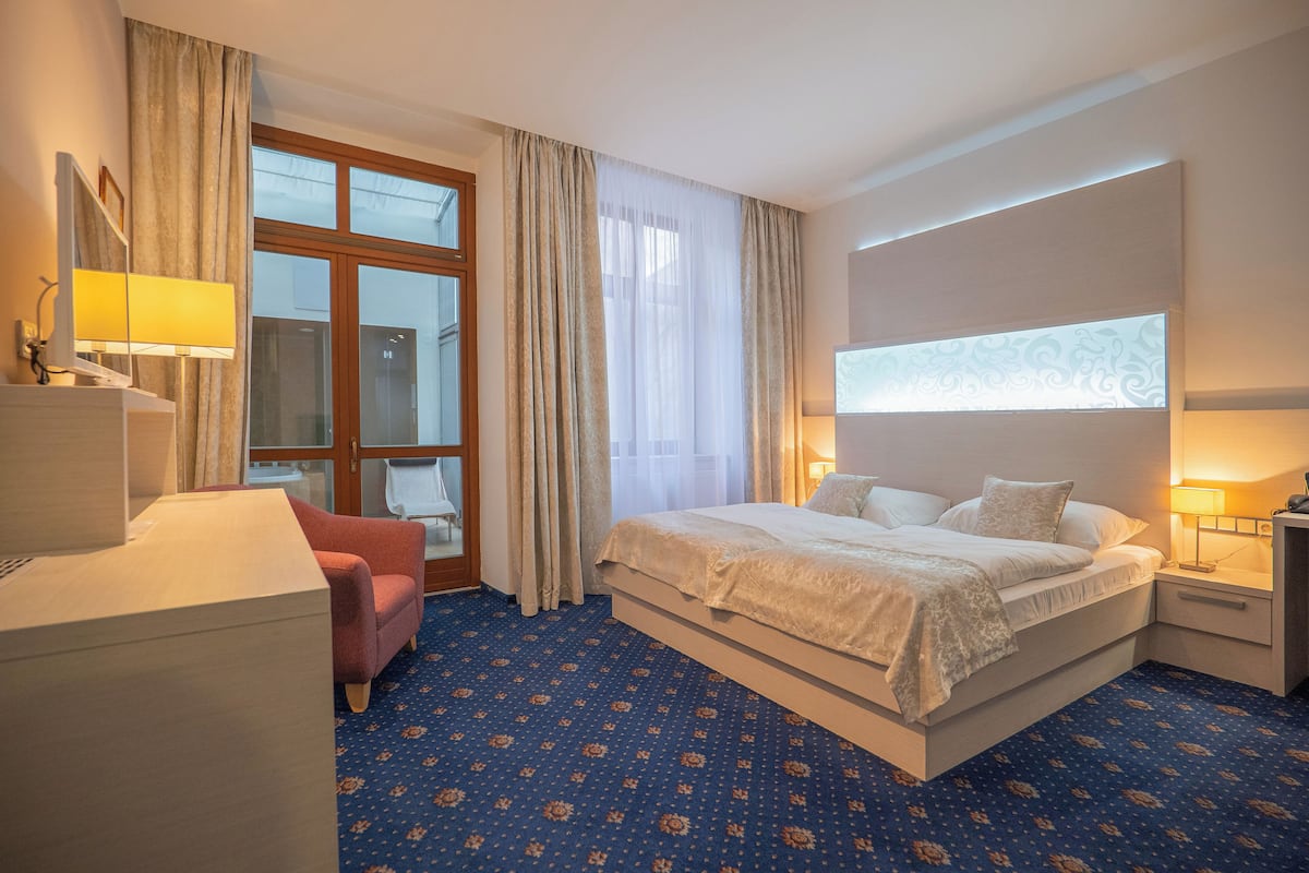 A hotel room with a large bed, a desk, a chair, a window with curtains, and a patterned carpet.