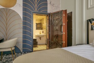 Habitación de hotel con cama, una silla, baño con lavamanos y una puerta de cristal que da a la zona de ducha.