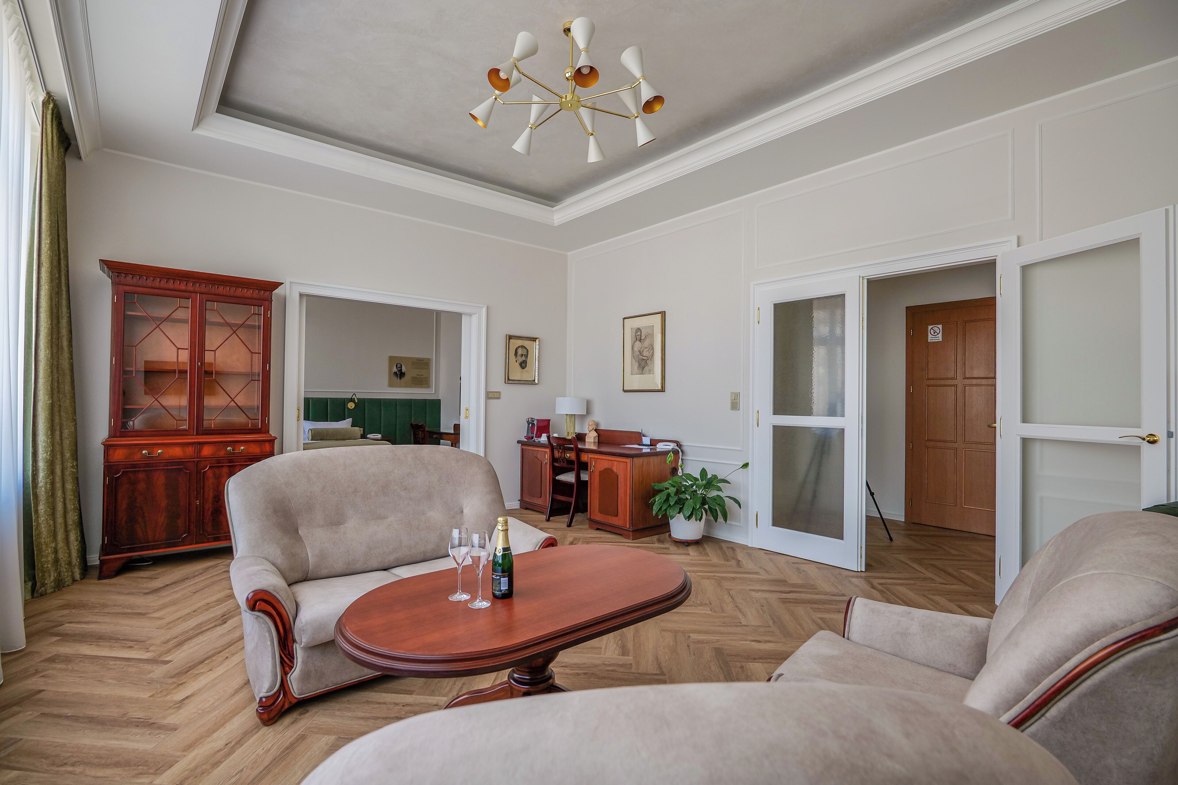 A spacious living room with a wooden floor, a large armchair, a coffee table with a bottle and glasses, a wooden cabinet, and a ceiling light fixture.
