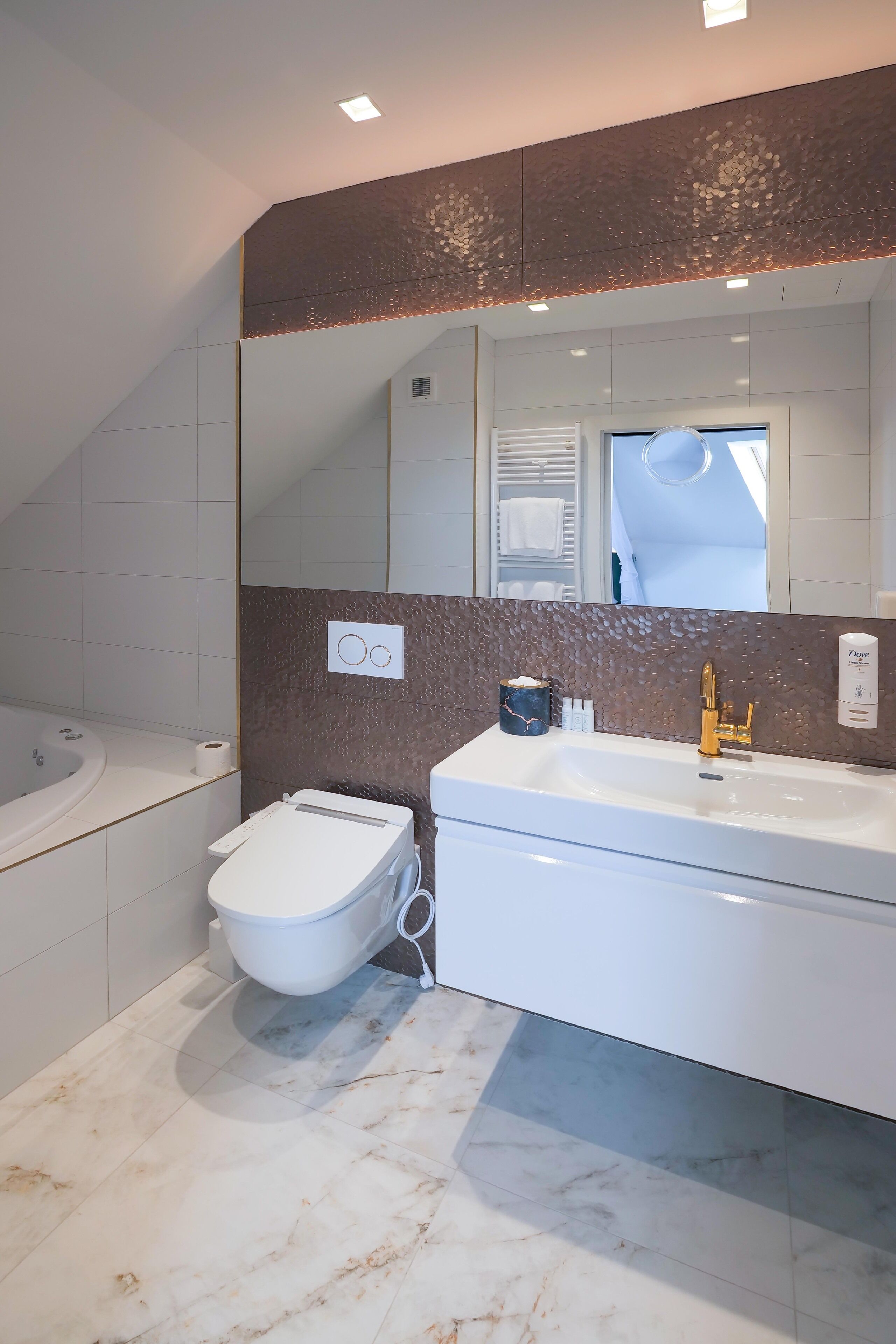 A modern bathroom with a white bathtub, a toilet, and a sink with a gold faucet. There is a large mirror above the sink and a textured wall behind it.