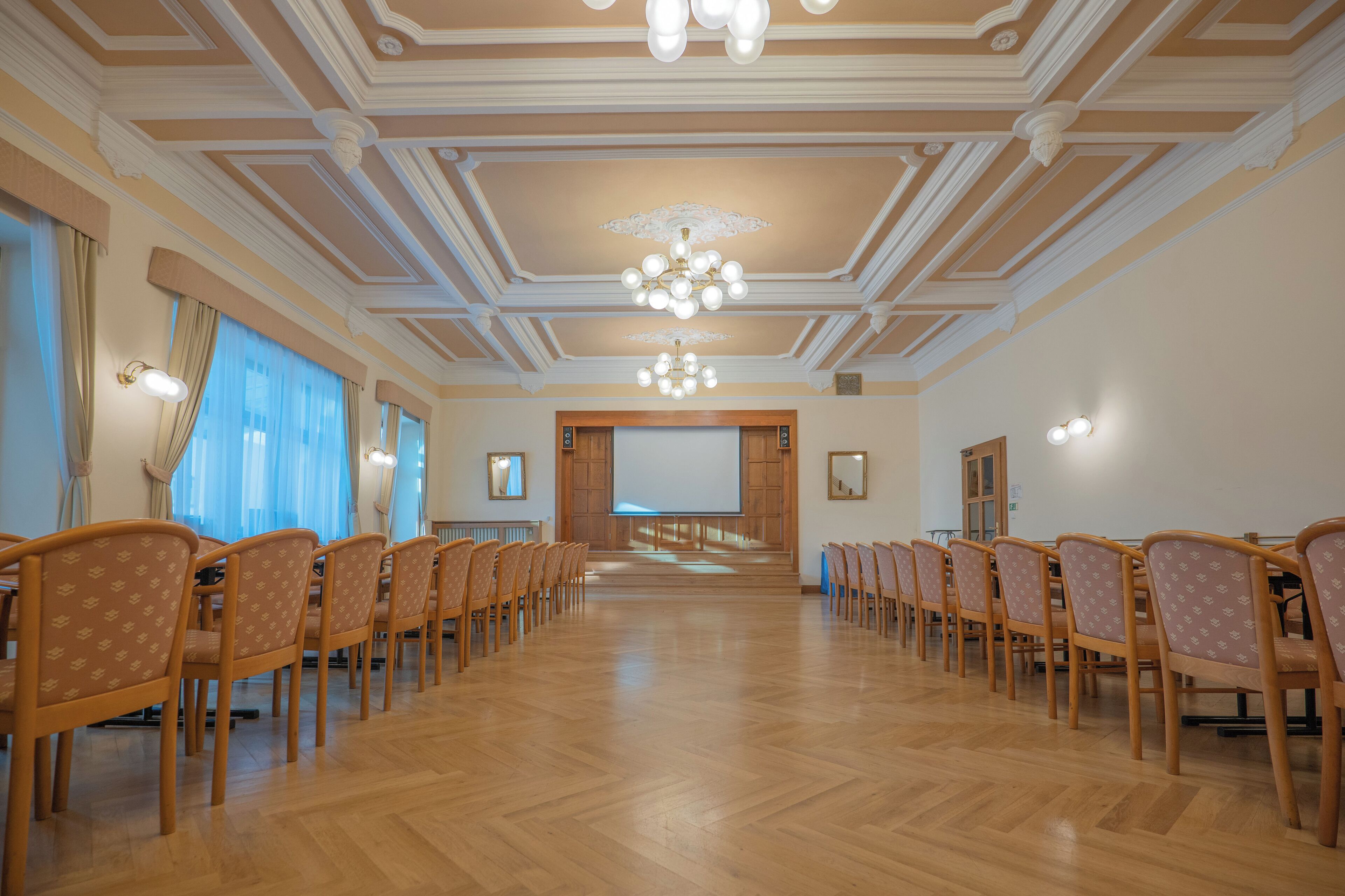 Un salón de conferencias con filas de sillas de madera, una pantalla grande al frente y un candelabro.