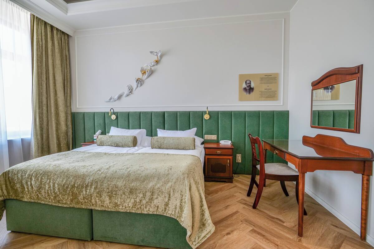 A neatly arranged bedroom with a green upholstered headboard, a wooden nightstand, a desk with a chair, and a wall-mounted telephone.