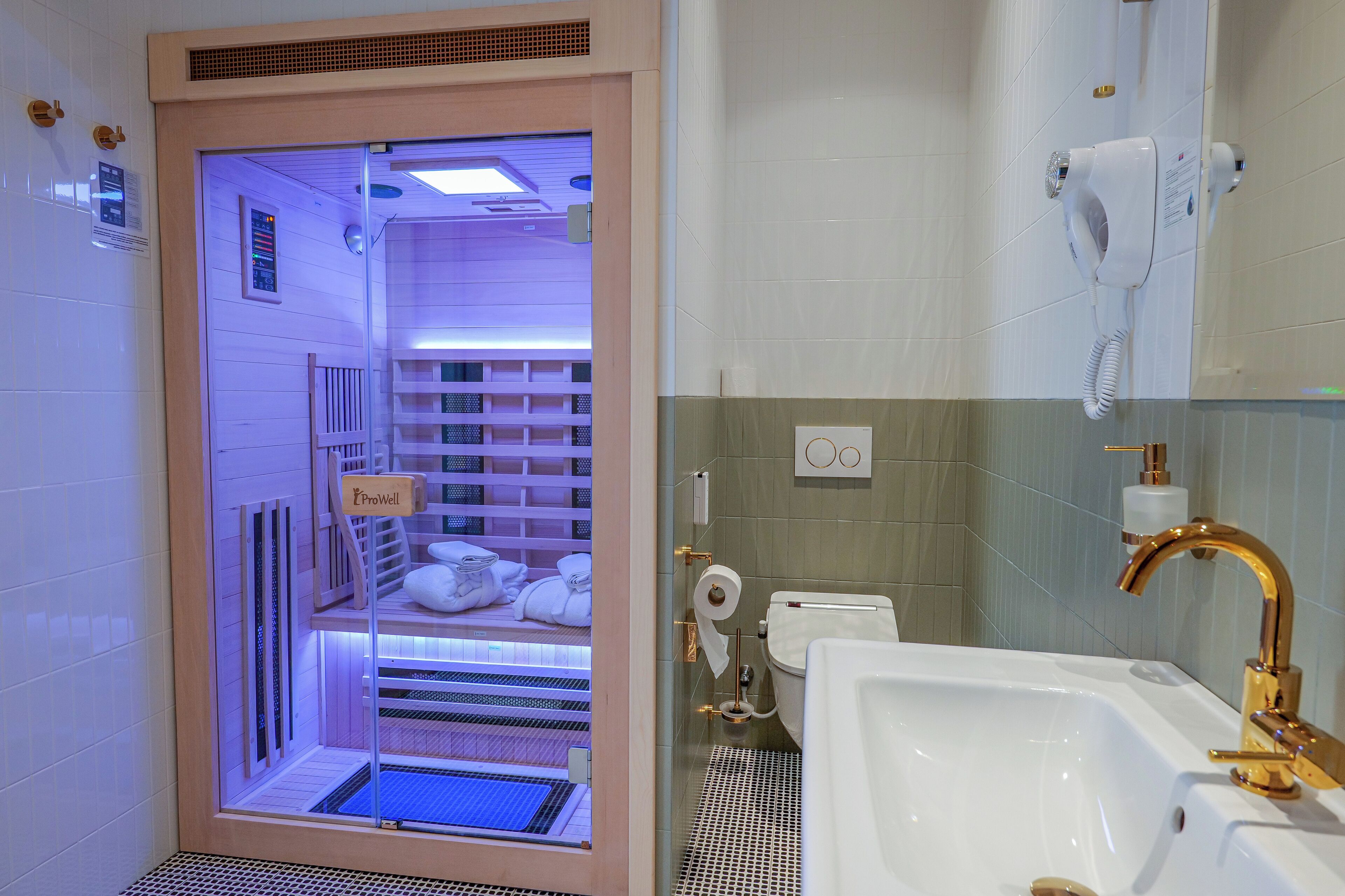A modern bathroom with a built-in sauna, a white sink with gold fixtures, and a ProWell sauna visible through a glass door.