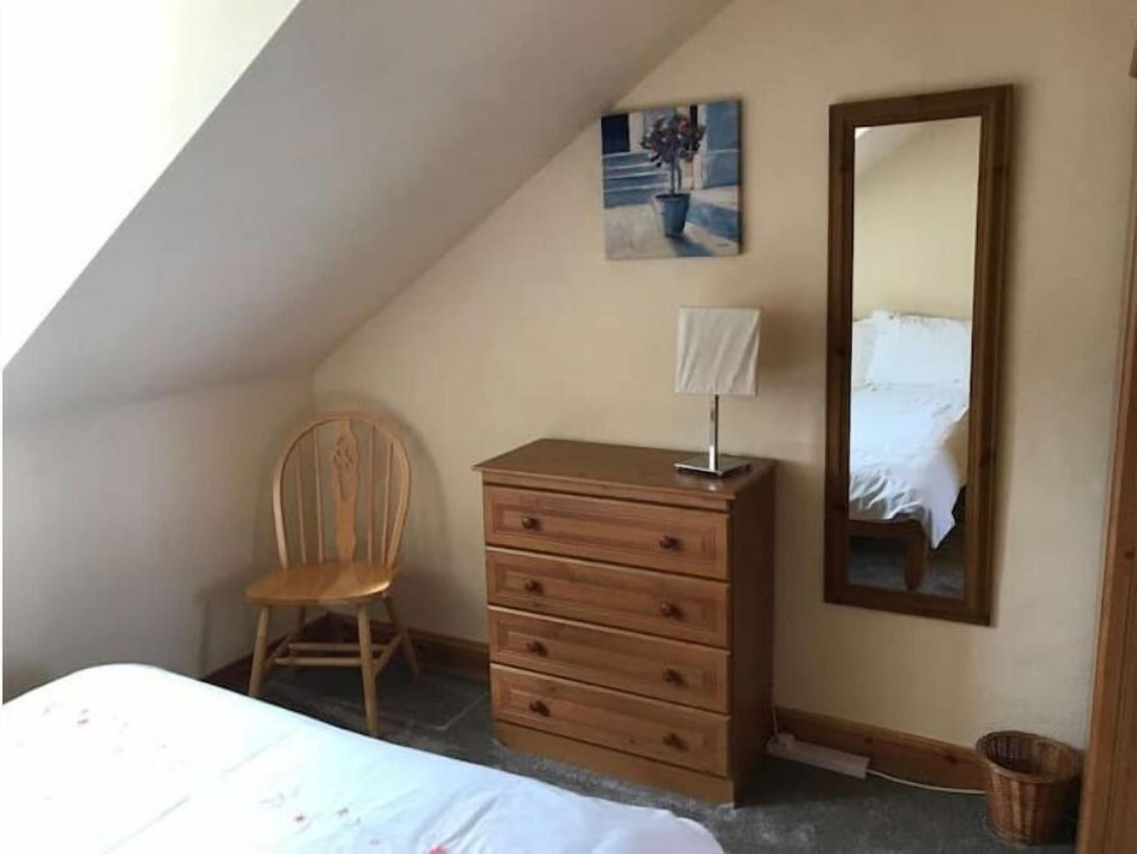 A bedroom with a bed, a wooden dresser, a chair, a painting, and a mirror.