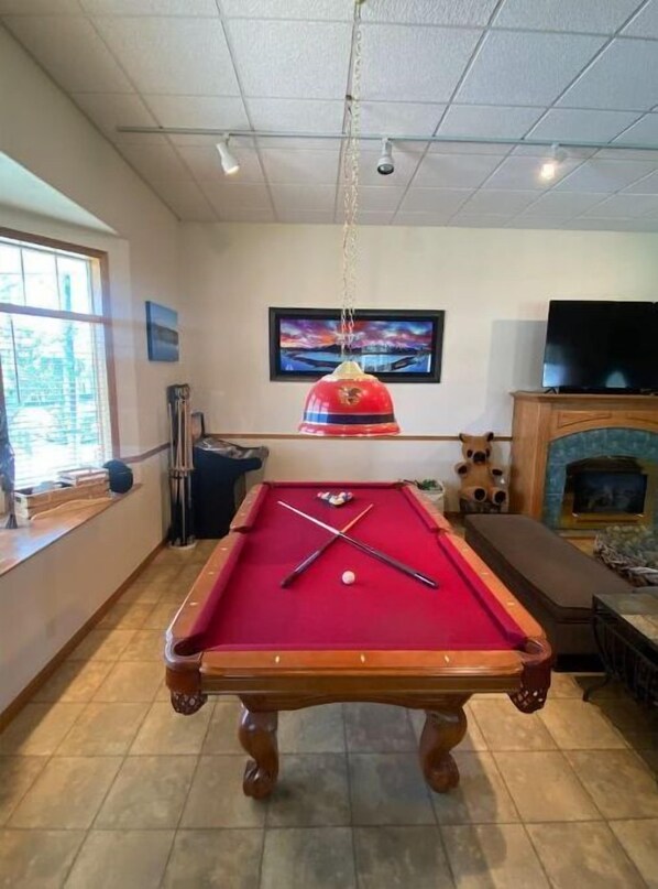 Una mesa de billar en una habitación con chimenea, un televisor en la pared y una ventana con persianas.