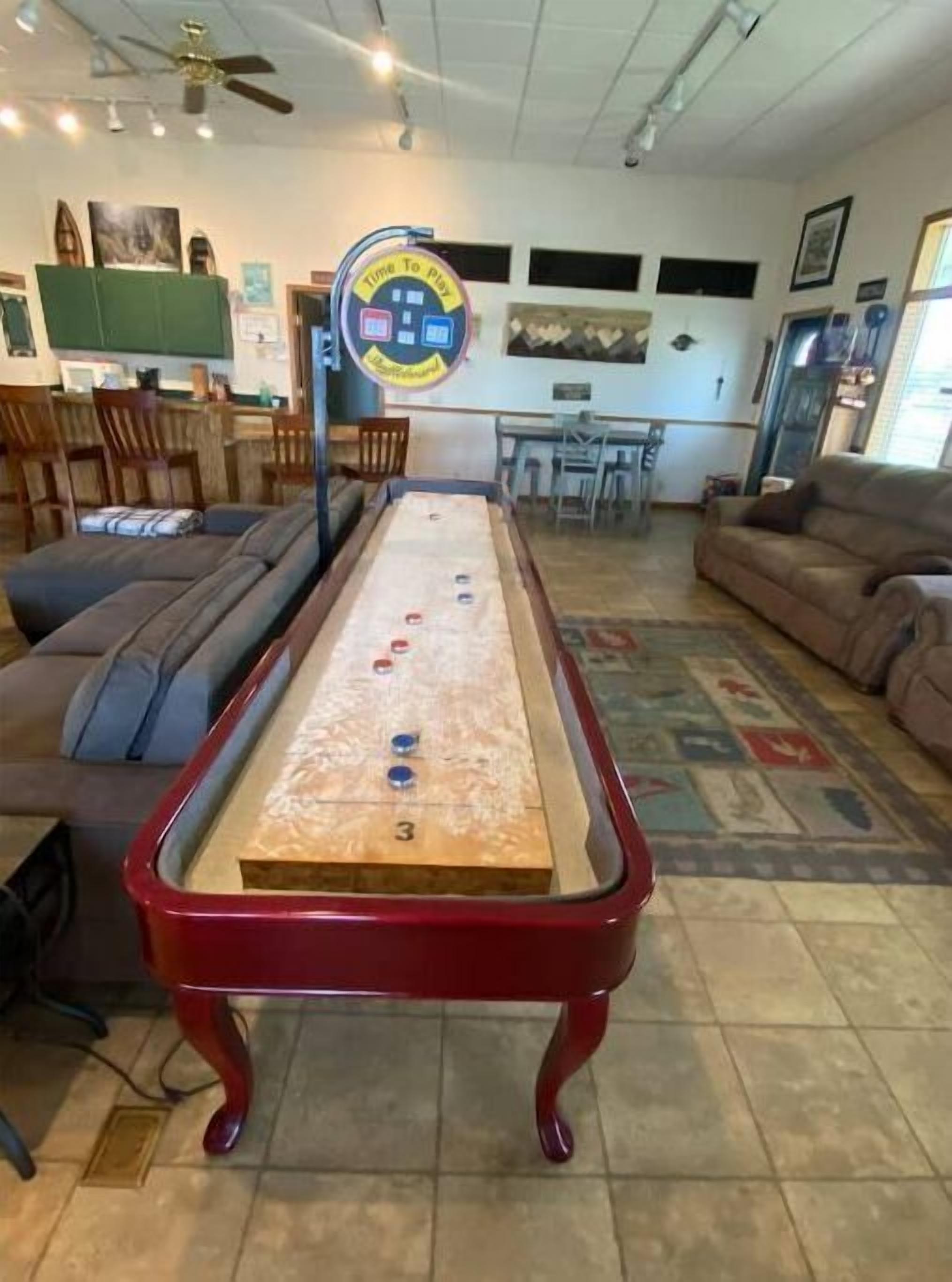 A shuffleboard table in a living room with a green couch, a brown sofa, and a dining area in the background.