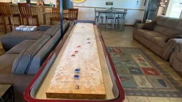A shuffleboard table in a living room with a green couch, a brown sofa, and a dining area in the background.