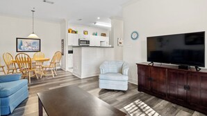 A modern living room with a TV, a dining area with chairs, a sofa, and a kitchen in the background. - Ball fields! OWA! & Near the Beach! (Foley)