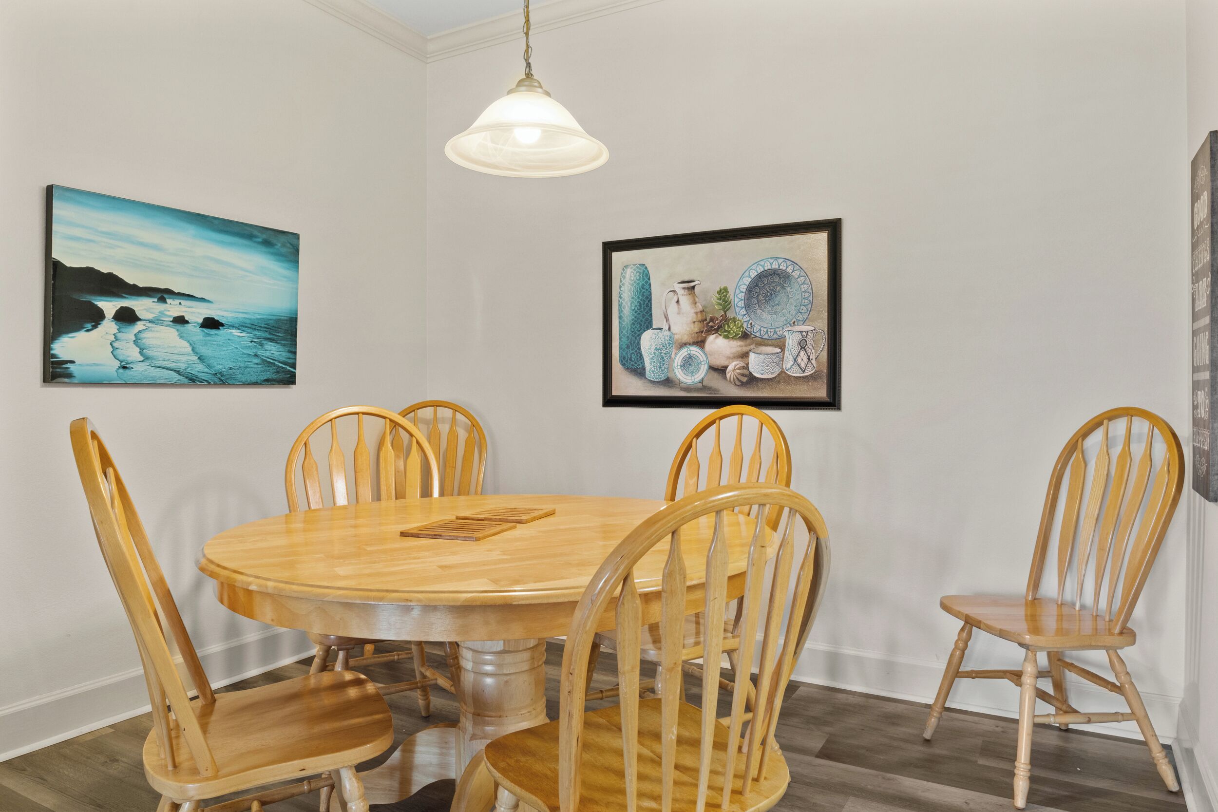 A dining area with a round wooden table and six wooden chairs, surrounded by white walls and two framed artworks.