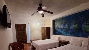 A room with two beds, a ceiling fan, a painting, a wooden wardrobe, and a television. - Casa Maru Hotel (Parras)