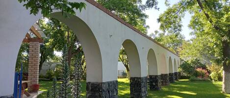 Un puente de piedra con arcos, rodeado de vegetación y un edificio blanco.