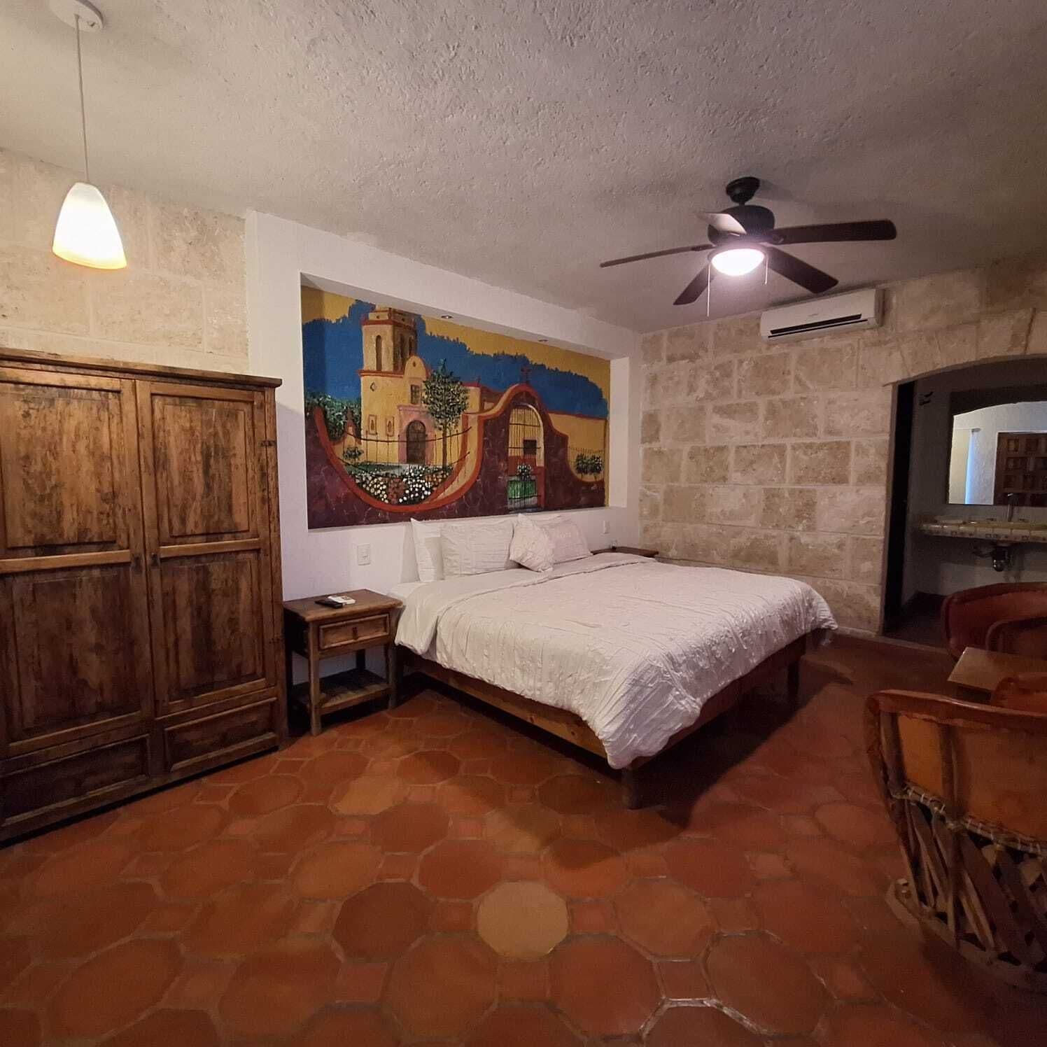 Une chambre à coucher avec un lit, une armoire en bois, un tableau au mur, un ventilateur de plafond et un sol carrelé.
