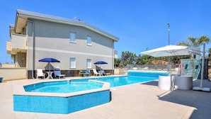 Un espace piscine comprenant deux piscines, des chaises longues, des parasols et un bâtiment doté d’un toit en tuiles.