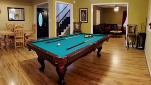 A pool table in a room with wooden flooring, a dining area with chairs, and a staircase.