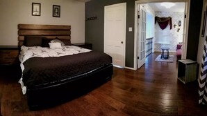 A bedroom with a wooden bed, a nightstand, a door leading to a bathroom, and a hallway with a chair and a small table.