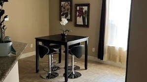 Een eetgedeelte met een tafel en vier barkrukken, een kroonluchter, bloemrijk kunstwerk in een lijst aan de muur, en een raam met gordijnen.