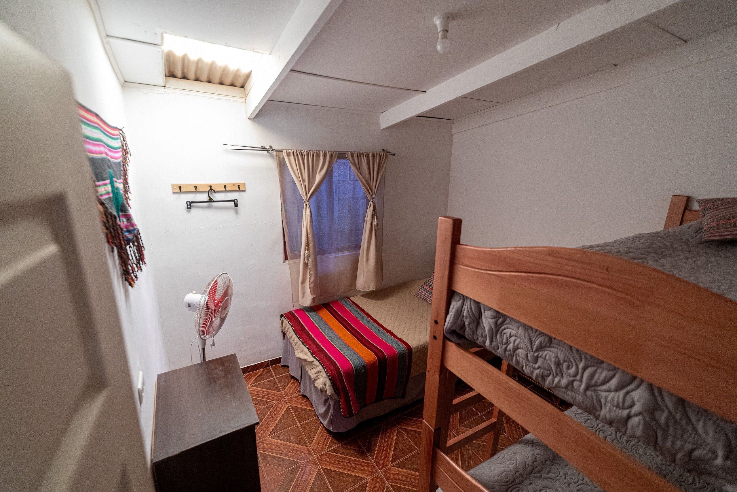 A dormitory room with a bunk bed, a small desk, a chair, a fan, and a window with curtains.