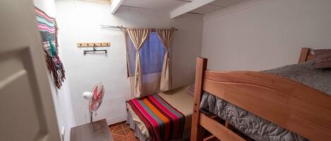 A dormitory room with a bunk bed, a small desk, a chair, a fan, and a window with curtains.