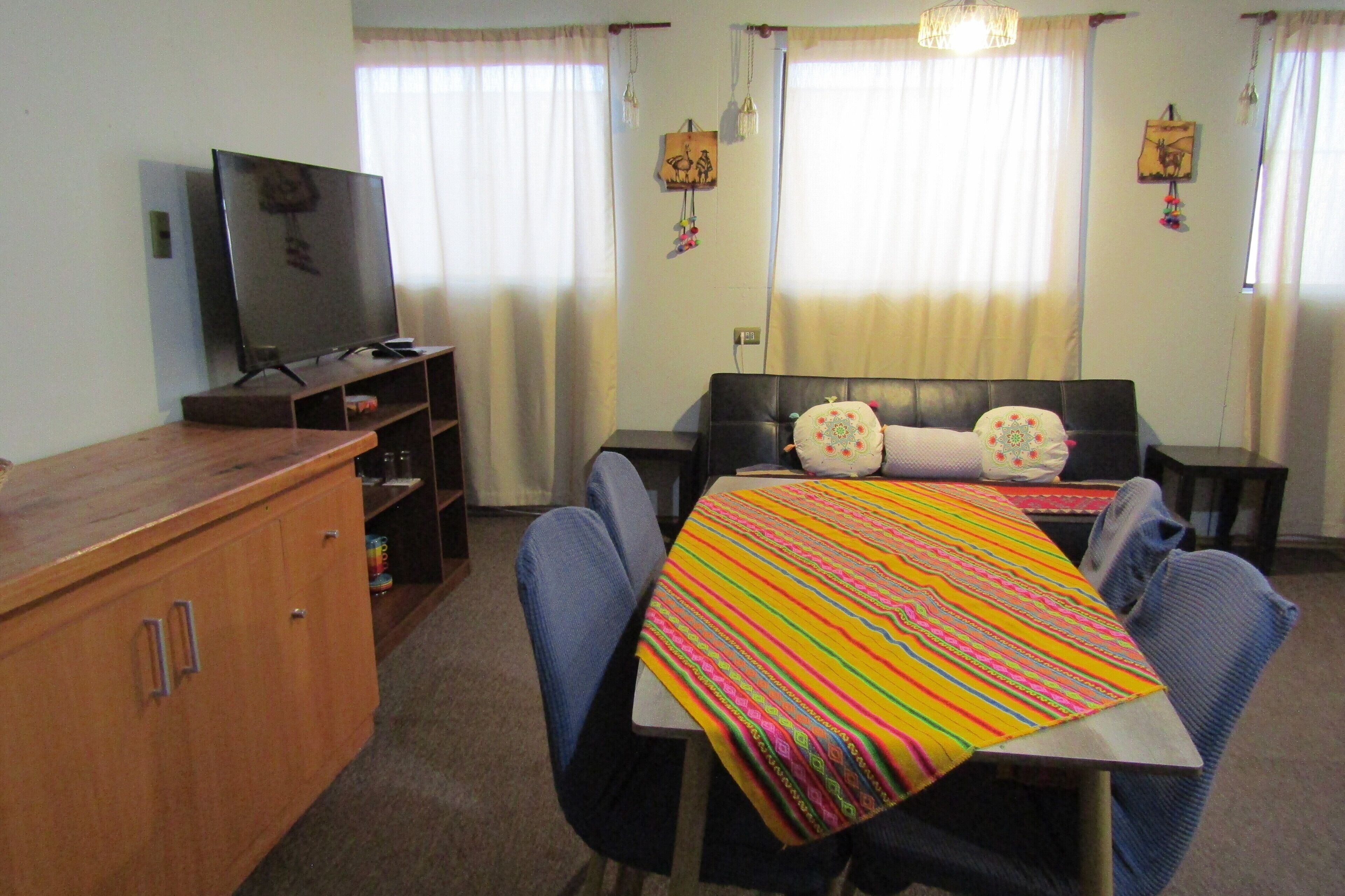 A living room with a television, a sofa, a dining table with chairs, and a colorful blanket.