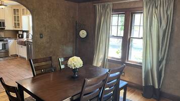 A dining room with a wooden table, chairs, and a vase of flowers. There is a kitchen in the background with cabinets and appliances.