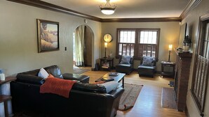 A living room with a black leather sofa, a coffee table, and a fireplace. There is a clock on the wall and a window with curtains. - Historic Twin Tudors Inn Southwest Room (KP1404SW) (Sioux Falls)