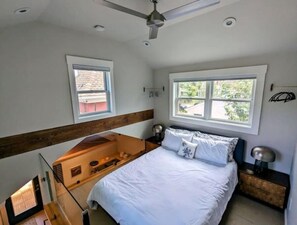 A bedroom with a bed, a window, a ceiling fan, and a small table with a lamp.