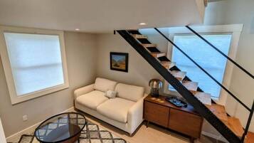 Un salon comprenant un canapé, une table basse en verre, un tapis à motifs, un tableau accroché au mur et un escalier avec une rampe en métal.