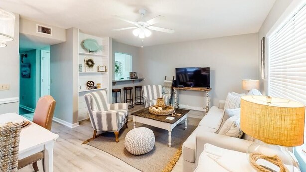 Un salon comprenant un canapé, des fauteuils, une table basse et un téléviseur. On y trouve également un ventilateur de plafond, un téléviseur fixé au mur et un coin repas avec une table et des chaises.