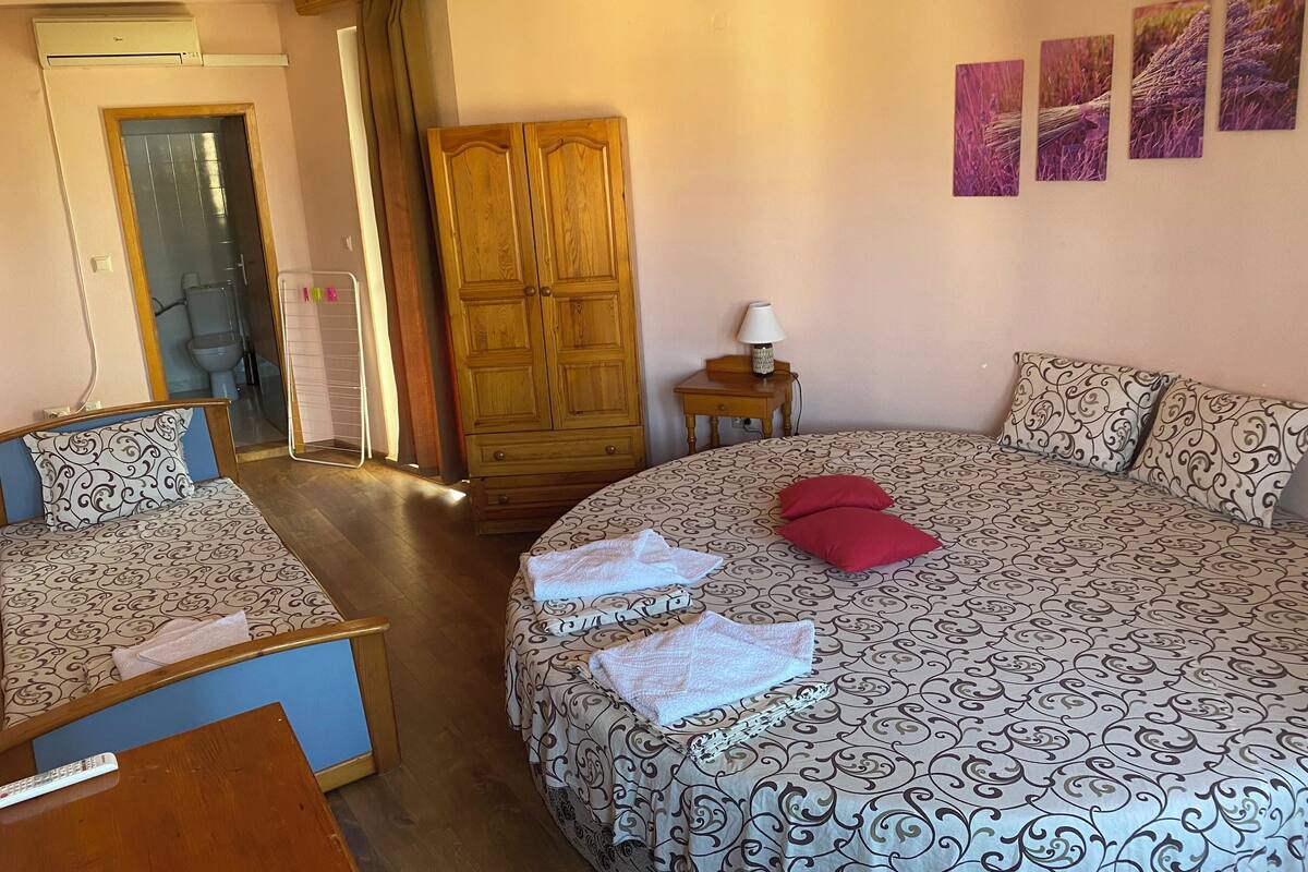 A bedroom with two beds, a wooden wardrobe, a nightstand with a lamp, and a bathroom visible through a glass door.