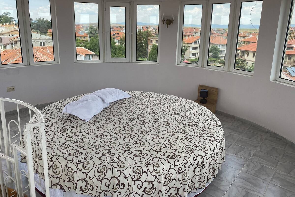 A bedroom with a large bed, multiple windows, and a view of the cityscape.