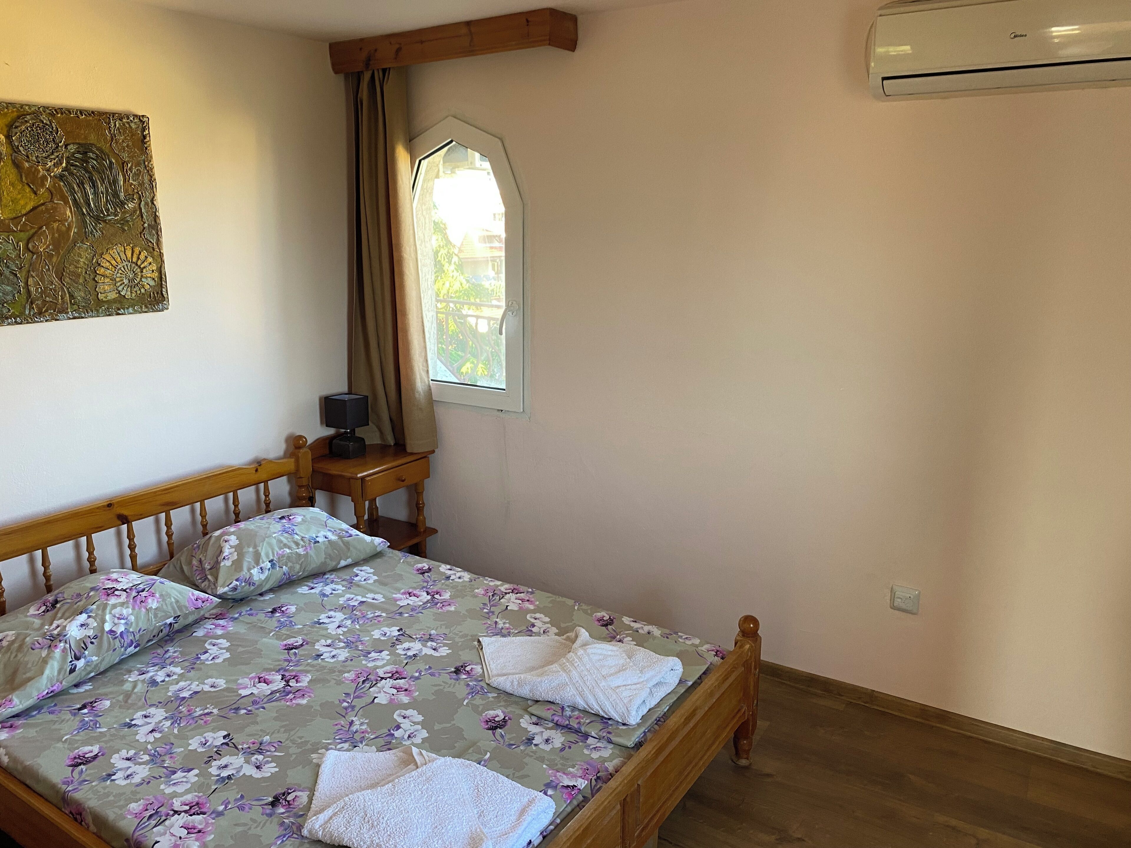 A bedroom with a bed, a nightstand, a lamp, a window with curtains, and an air conditioner.