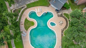 Vista aérea de uma piscina de formato peculiar com hidromassagem, cercada por um gramado bem cuidado e um edifício de tijolos ao fundo.