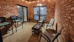 Um espaço de pátio com paredes de tijolo, assentos, uma porta de vidro que dá para um espaço interno, e teto com vigas e iluminação aparentes.