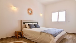 Quarto com cama, um banquinho de madeira, tapete redondo, luminária fixada na parede e uma janela.