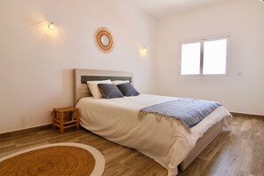 Ein Schlafzimmer mit einem Bett, einem kleinen Holzschemel, einem runden Teppich, einer Wandleuchte und einem Fenster.