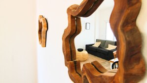 Um espelho reflete um quarto com um sofá preto, um banquinho redondo de vime e uma mesa pequena.