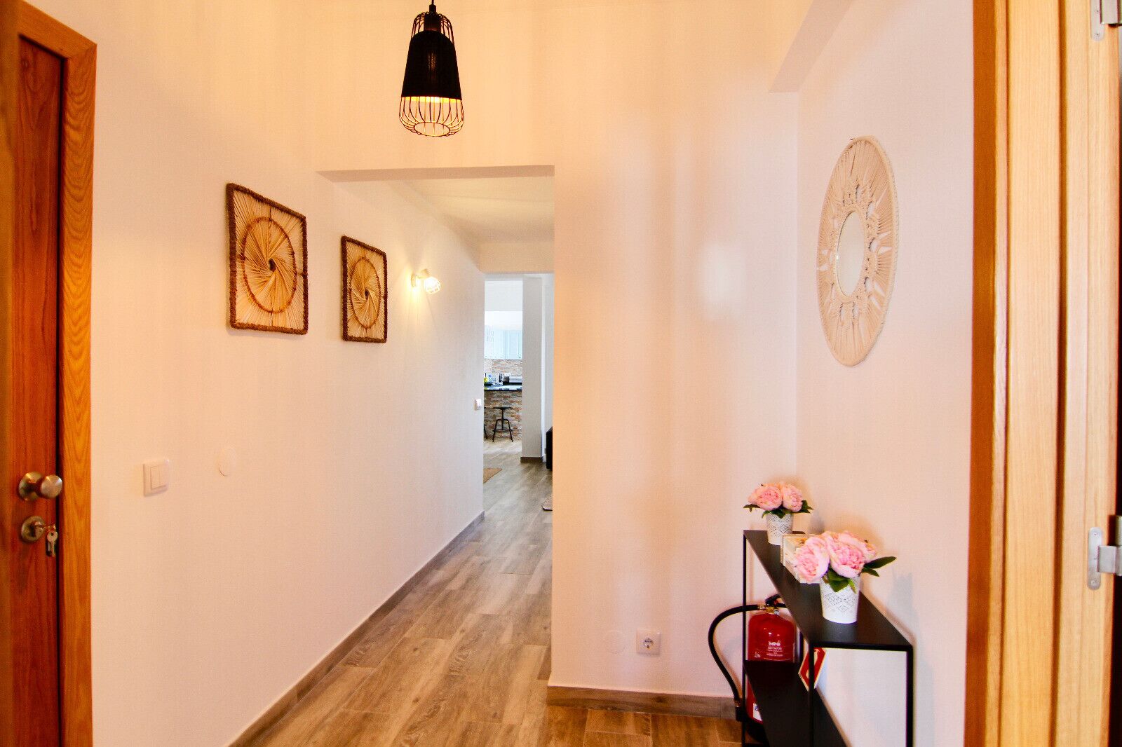 Un pasillo con piso de madera, una mesa negra con flores rosadas y un reloj de pared.