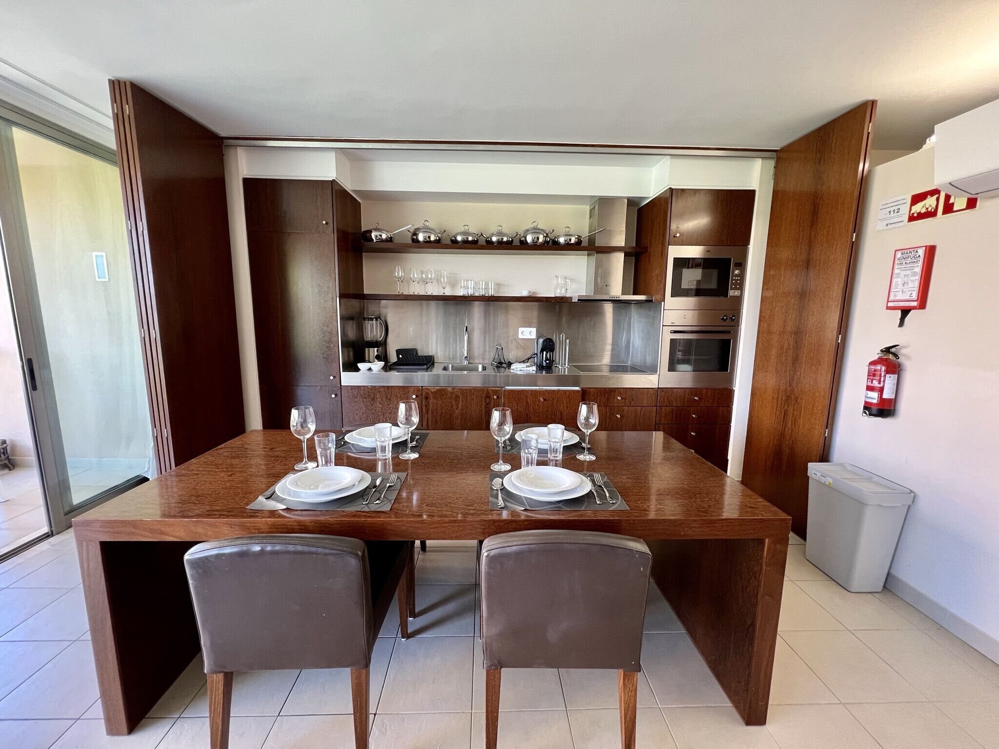 A modern kitchen with stainless steel appliances, a dining table set for four, and a fire extinguisher on the wall.