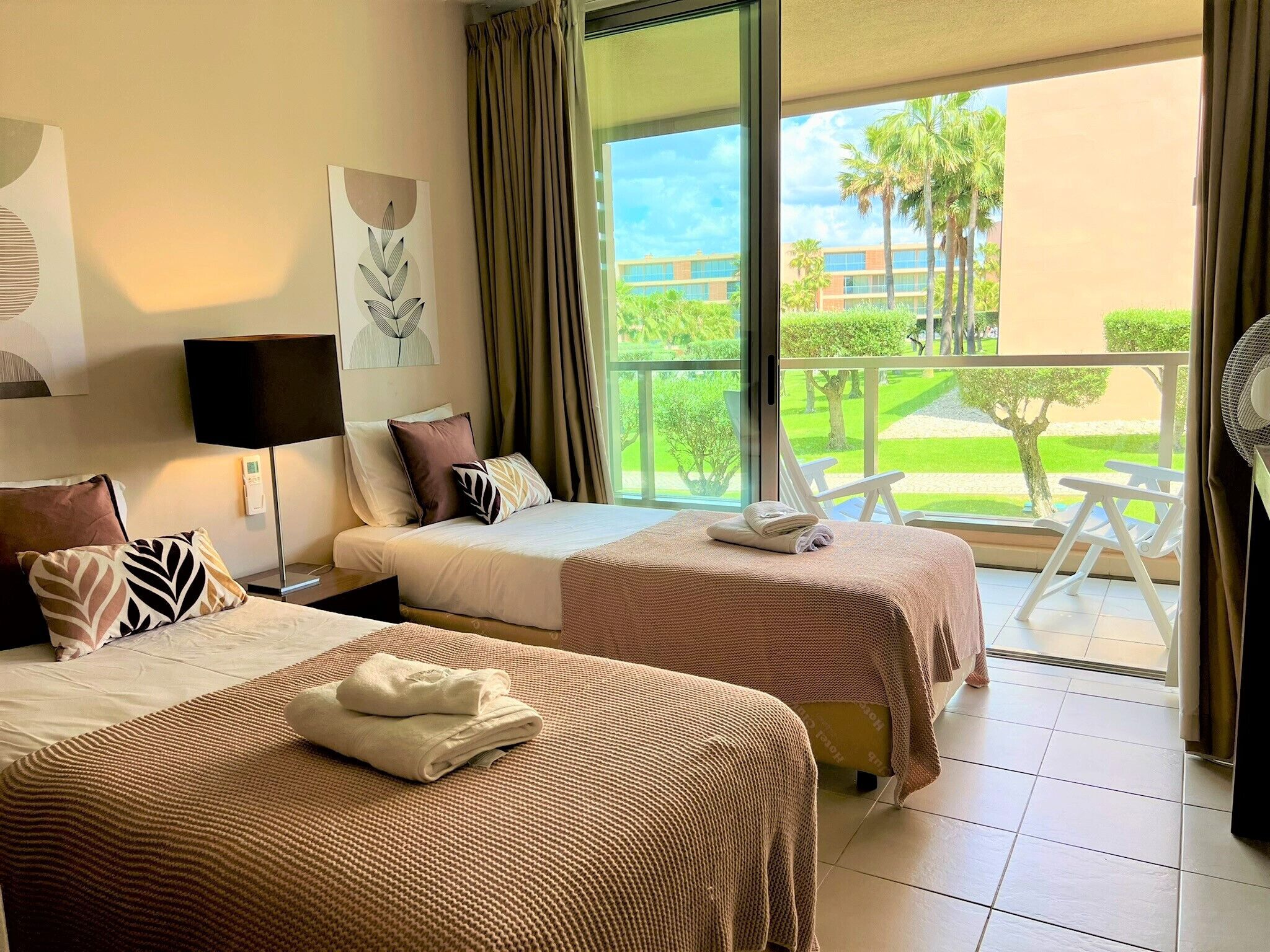 Habitación de hotel con dos camas, balcón y vista a un edificio y follaje.