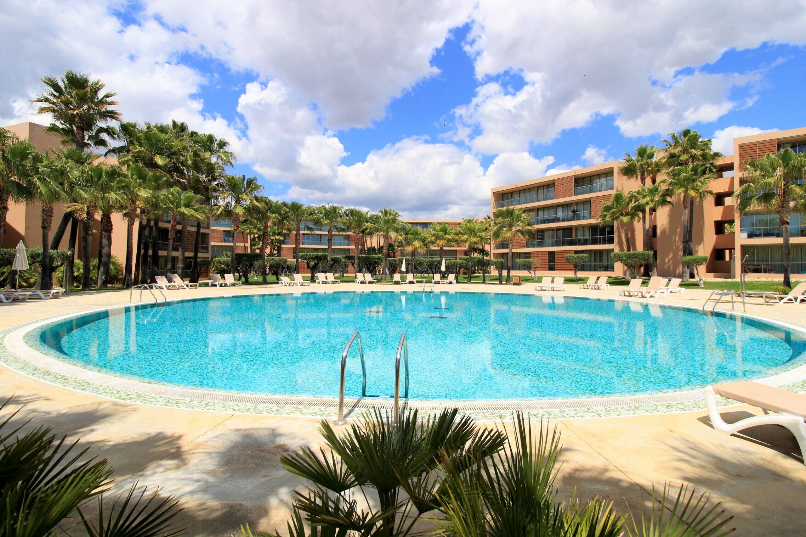 A large, clear swimming pool surrounded by lounge chairs and palm trees, with modern buildings in the background.