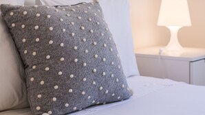 A neatly made bed with a grey polka-dotted pillow and white pillows, a white bedside table with a lamp, and a white wall. - Lagos Classic by Homing (Lagos)