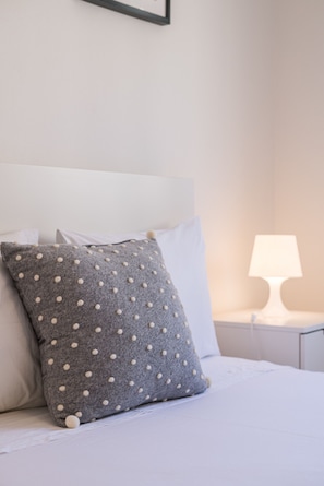 Ein ordentlich bezogenes Bett mit einem grauen Kissen mit Punkten und weißen Kissen, ein weißer Nachttisch mit einer Lampe sowie eine weiße Wand.
