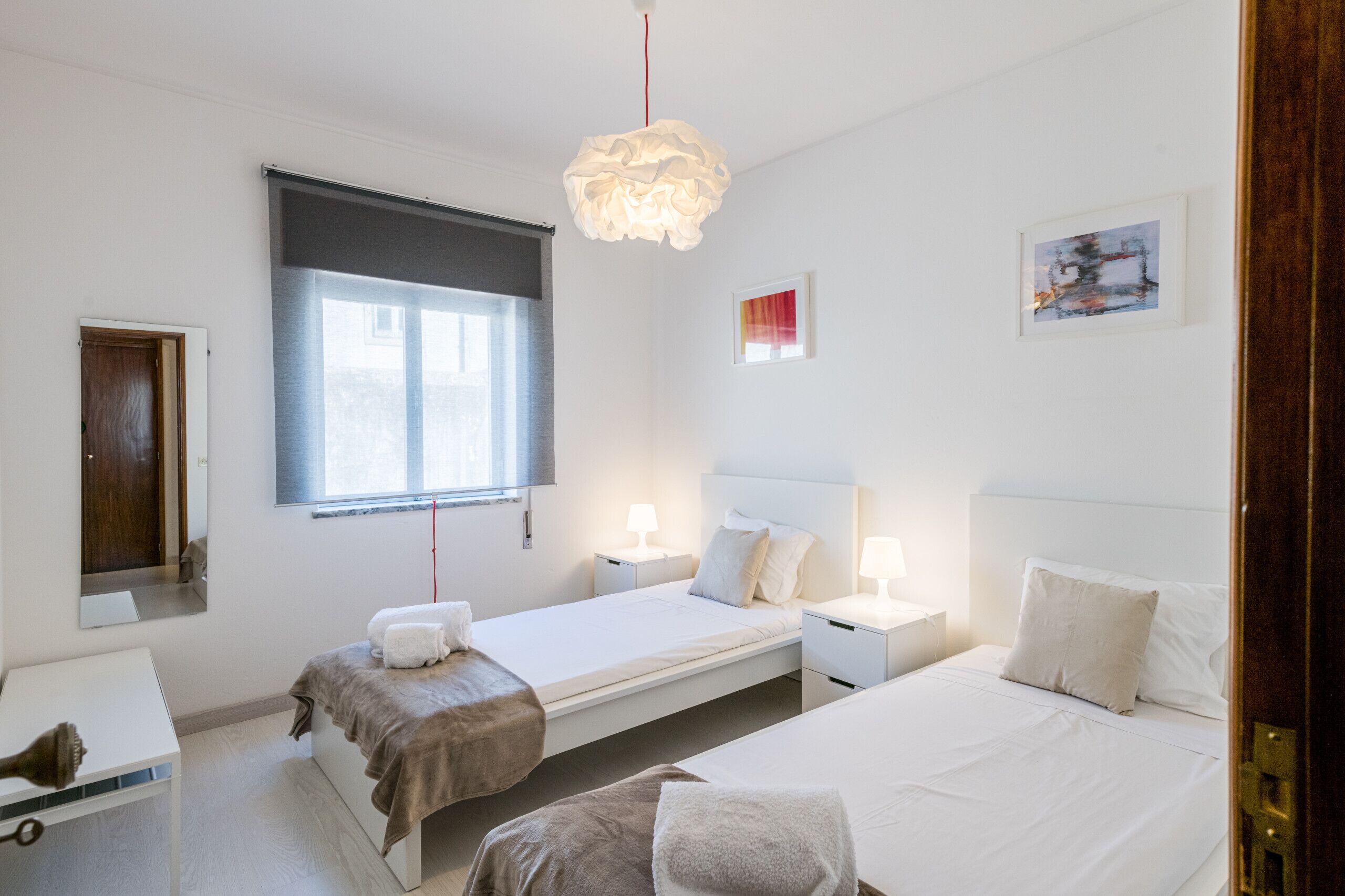 A bedroom with two beds, a window, a bedside table with a lamp, and a wall-mounted artwork.